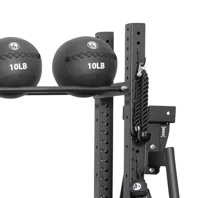 Two black 10LB medicine balls sit on a rack featuring the Bells of Steel Utility Hook. A thick black rope and the All-in-One Trainer frame with numbered height adjustment holes complete this versatile gym storage setup.