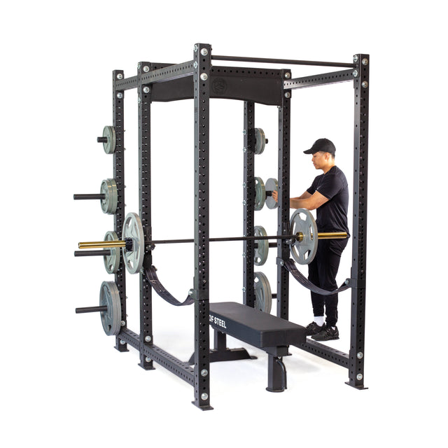 A person in black workout clothes and a cap stands inside the Bells of Steel Hydra Six Post Power Rack - Prebuilt (3" x 3", ⅝" Holes) with weight plate storage, a loaded barbell, and bench in their home gym, ready to exercise.