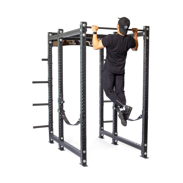 A person in black athletic wear and a cap does a pull-up on the Bells of Steel Hydra Six Post Power Rack - Prebuilt (3" x 3", ⅝" Holes) with weight plate storage, ideal for any home gym, against a white background.