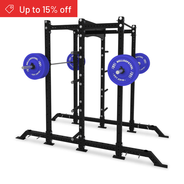 A black Bells of Steel Hydra 2-Sided Rack power rack holds a barbell with blue plates. A red "Up to 15% off" banner is in the top left corner. The background is plain white.