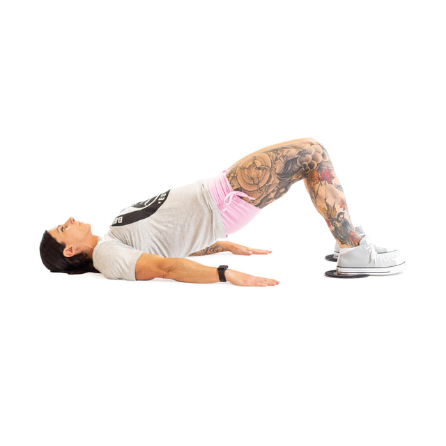 A tattooed individual in a gray t-shirt, pink shorts, and sneakers uses Bells of Steel Core Sliders for a glute bridge, lifting hips off the floor—an effective total body exercise.