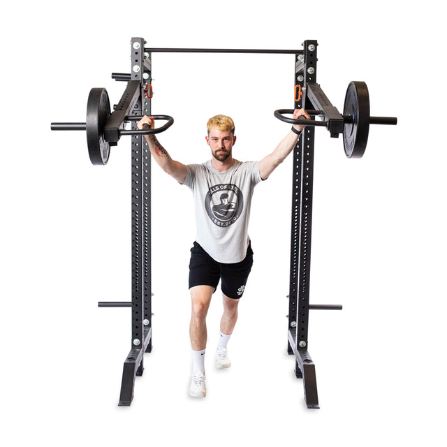A man lunges while gripping the Bells of Steel Lever Arms Rack Attachment, loaded with Olympic weight plates, in a gym. He wears a light gray t-shirt, black shorts, and white sneakers against a plain white background.