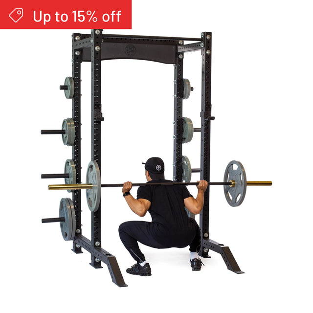 A person in black workout gear squats with a loaded barbell inside the Bells of Steel Hydra Collegiate Power Rack Builder (3" x 3", ⅝" Holes), with weight plates stored on the rack. A red banner says "Up to 15% off.