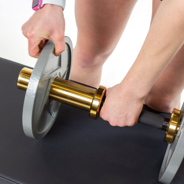 A person attaches a Bells of Steel Loadable Dumbbell Handle to a gold and black barbell, wearing a smartwatch and standing over a black bench amidst home gym equipment, preparing for a workout.