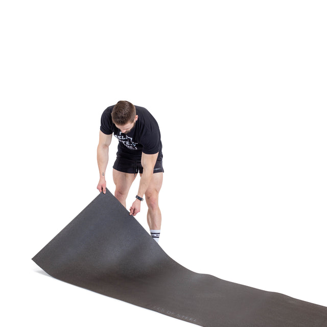 A person stands on a white background, unrolling a large black Bells of Steel Cardio Machine Floor Mat while wearing a black t-shirt, black shorts, and socks.