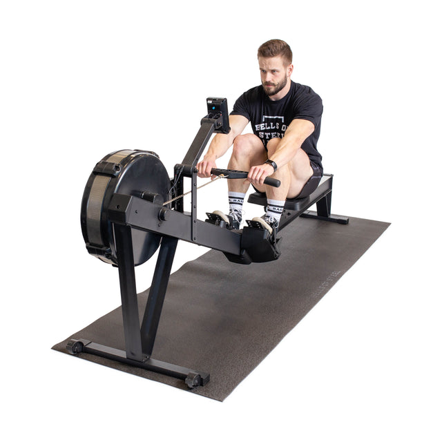 A man in athletic wear uses a rowing machine on a Bells of Steel Cardio Machine Floor Mat against a white background.