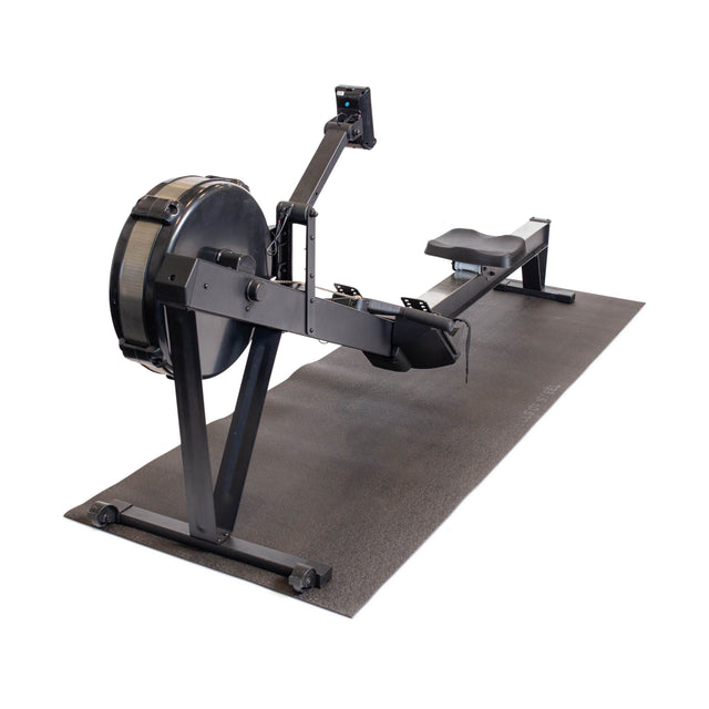 A black indoor rowing machine is placed on a Bells of Steel Cardio Machine Floor Mat, with a flywheel, sliding seat, footrests, and a monitor for workout tracking. The setup is pictured against a white background.