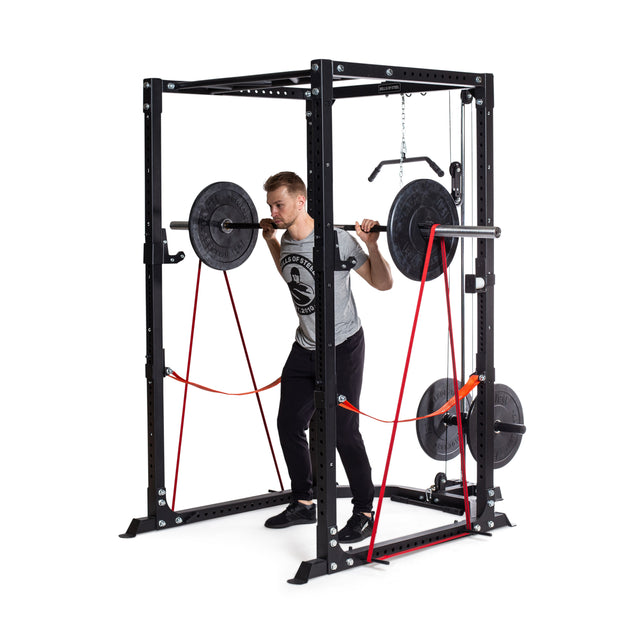A man performs a barbell squat inside a black power rack, using Bells of Steel 41" Fabric Non-Slip Resistance Bands and weight plates for powerlifting resistance, against a white background.