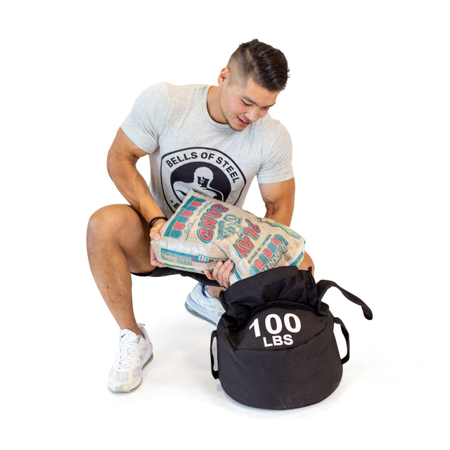 A man in athletic gear pours sand labeled "Bells of Steel" into a black Fitness Sandbags duffel marked "100 LBS," demonstrating the versatile Bells of Steel Fitness Sandbags for workouts, with a white background.