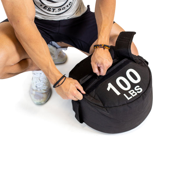 A person in athletic wear and white sneakers secures the strap of a black Bells of Steel Fitness Sandbag Set labeled "100 LBS," featuring durable Condura construction, against a white background.
