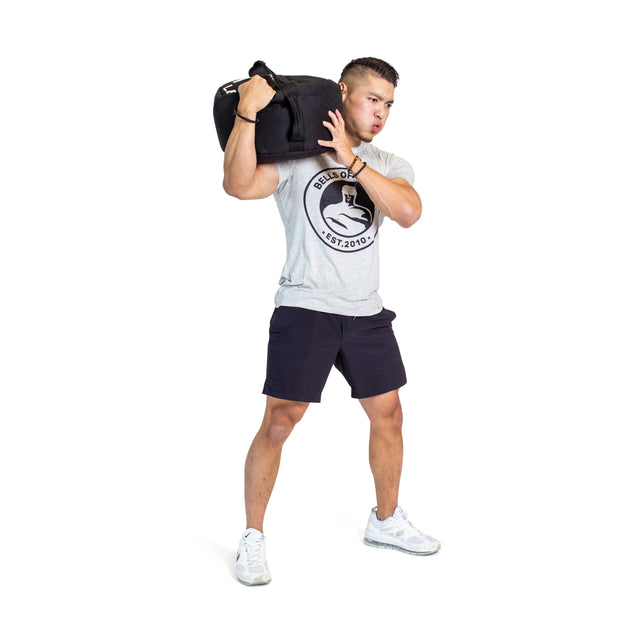 A man in a gray t-shirt and black shorts lifts a Bells of Steel Fitness Sandbag Set onto his right shoulder, demonstrating sandbag exercises with bent knees against a plain white background.