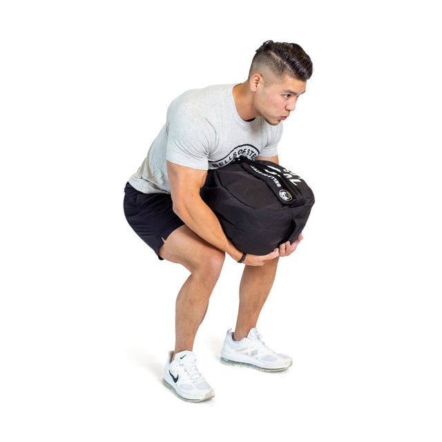 A man in a gray t-shirt, black shorts, and white sneakers squats and lifts a Bells of Steel Fitness Sandbag Set with his back straight and arms holding the bag in front of him against a white background.