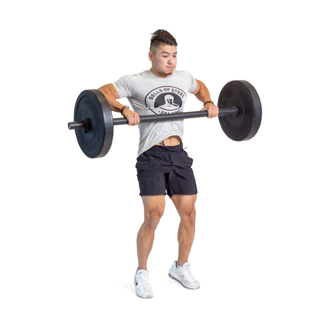 Wearing a grey t-shirt, black shorts, and white sneakers, a person performs an upright row with the Bells of Steel Axle Bar, lifting it to chest height and testing their grip strength against a white background.