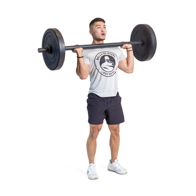 A man in a gray Bells of Steel t-shirt and black shorts stands on a white background, gripping Axle Bars at his shoulders with a focused look, ready to test his grip strength.