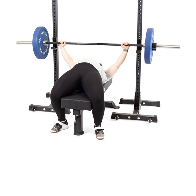 A person bench presses a barbell with blue plates, using Bells of Steel Squat Stands (2.3" x 2.3", ⅝" holes), while wearing a white shirt, black leggings, and athletic shoes—an ideal setup for any home gym.
