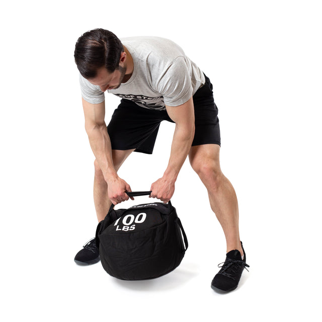 A man in a gray T-shirt and black shorts bends over, gripping the handle of a 100 lb Bells of Steel Fitness Sandbag Set made with durable Condura, preparing to lift it against a plain white background.