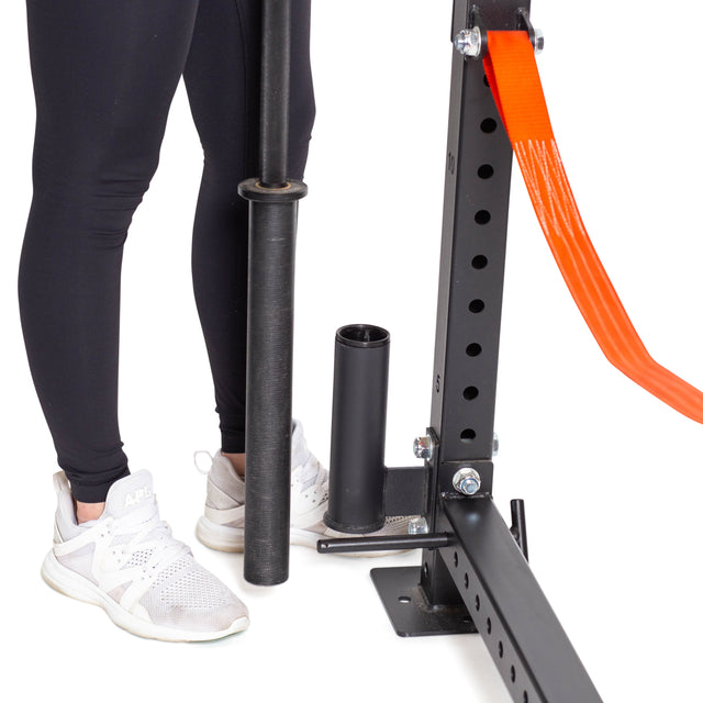 A person in black leggings and white sneakers stands next to a Bells of Steel Vertical Mount Barbell Holder Rack Attachment, efficiently storing a black barbell on a black metal gym rack with an orange safety strap.