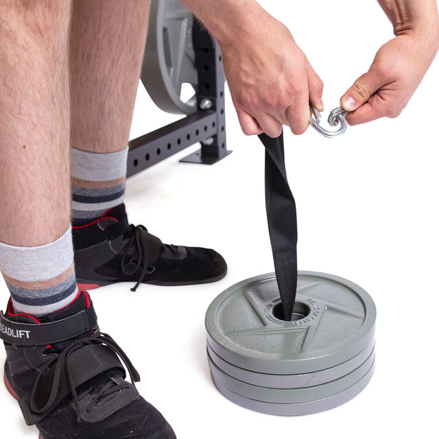 A user, with only their shoes and lower legs visible, attaches a strap with a carabiner through stacked weight plates to set up the Bells of Steel Cable Pulley for exercises.