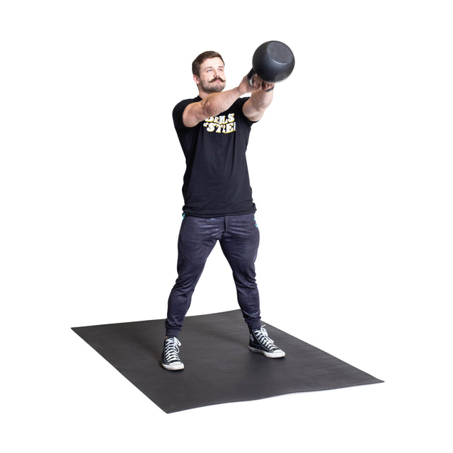 A man stands on Bells of Steel Premium Rubber Flooring 4' x 6', lifting a kettlebell at chest height. Wearing a black t-shirt, dark pants, and sneakers, he demonstrates gym floor protection during his workout.
