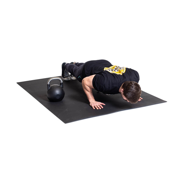 A man in a black T-shirt does push-ups on the Bells of Steel Premium Rubber Flooring 4' x 6', which offers excellent gym floor protection. A kettlebell sits beside him on the mat against a clean white background.