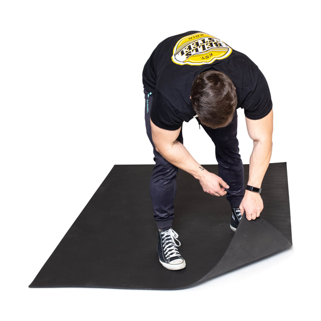 A person in a black shirt and dark pants bends down to lift the corner of a Bells of Steel Premium Rubber Flooring 4' x 6' mat, designed for gym floor protection.
