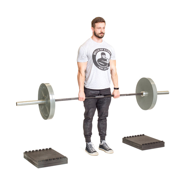 A man in a gray T-shirt and black pants deadlifts with a barbell. Nearby are Bells of Steel Stackable Pull Blocks, designed for space efficiency. The scene has a plain white background.
