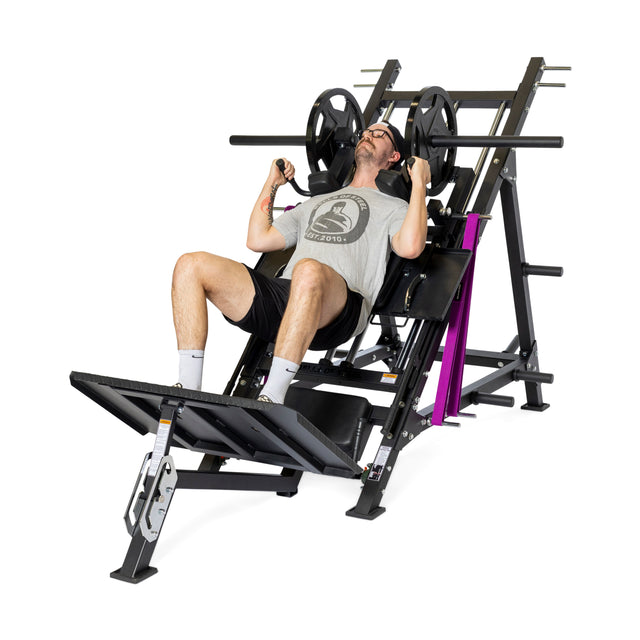 A person uses the Bells of Steel 2-in-1 Iso Leg Press & Hack Squat Machine (The Juggernaut), pushing the platform with their legs and gripping handles for support. They wear a gray t-shirt, black shorts, and white socks.