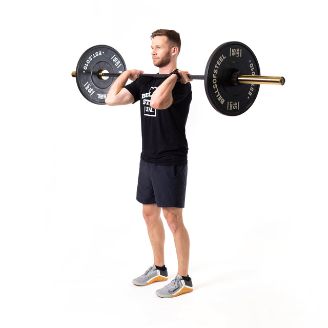A man, dressed in a black t-shirt, black shorts, and gray shoes, stands against a white background holding a Bells of Steel Olympic Weightlifting Barbell across his chest and shoulders, preparing to lift.