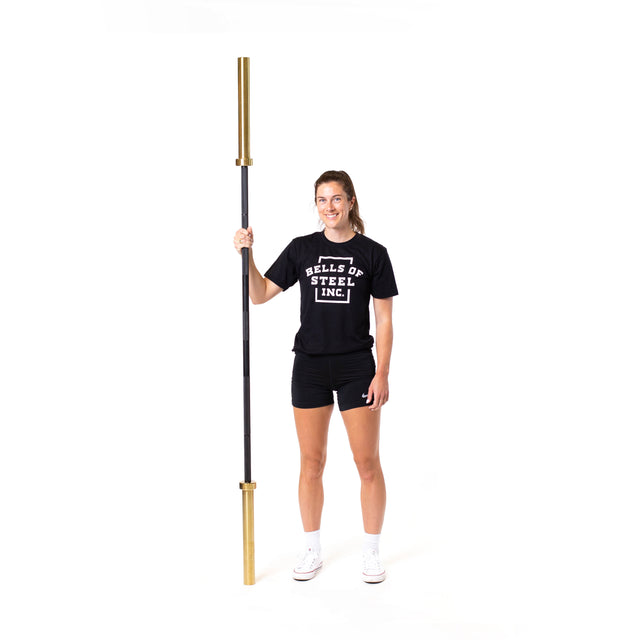 A person in a Bells of Steel t-shirt and shorts smiles while standing and holding a Bells of Steel Olympic Weightlifting Barbell vertically against a plain white background.