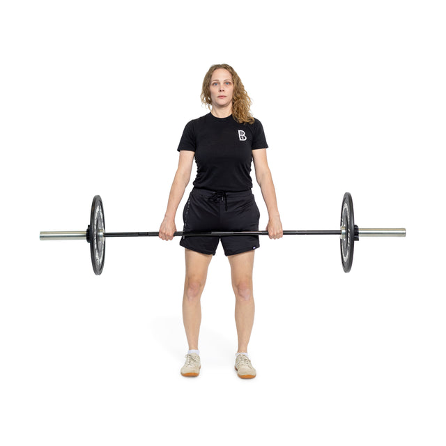 A woman stands on a white background, holding the Bells of Steel Juno Bar – Women’s Utility Bar with weights at thigh level. She is dressed in a black T-shirt, black shorts, and beige sneakers.