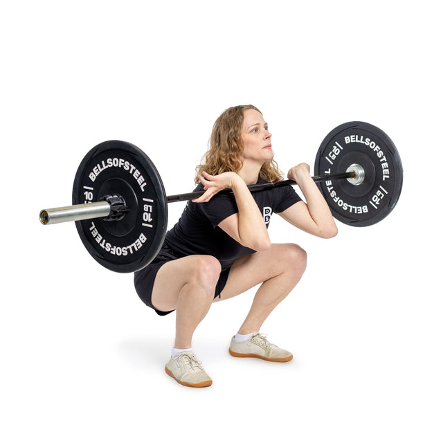 A woman with curly hair, dressed in black and beige shoes, performs a front squat with the Bells of Steel Juno Bar – Women’s Utility Bar on her shoulders against a white background.