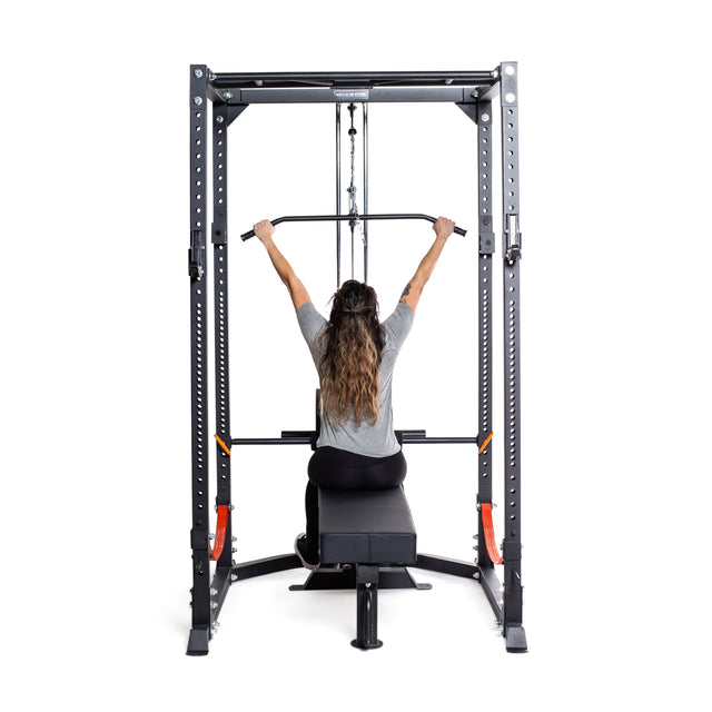 A person performs a lat pulldown using the Bells of Steel Lat Pulldown/Row Attachment. Seated on a bench in a gray shirt, they pull down the bar with both hands. The black machine with orange accents boasts an efficient pulley system for cable workouts.