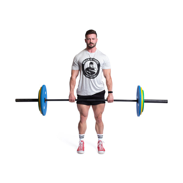 A man in a gray shirt and black shorts stands poised, clutching the Bells of Steel Calibrated Powerlifting Plates - LB Sets in blue and yellow. He wears red sneakers, has short hair and a beard, set against a plain white backdrop, perfectly embodying powerlifting precision.