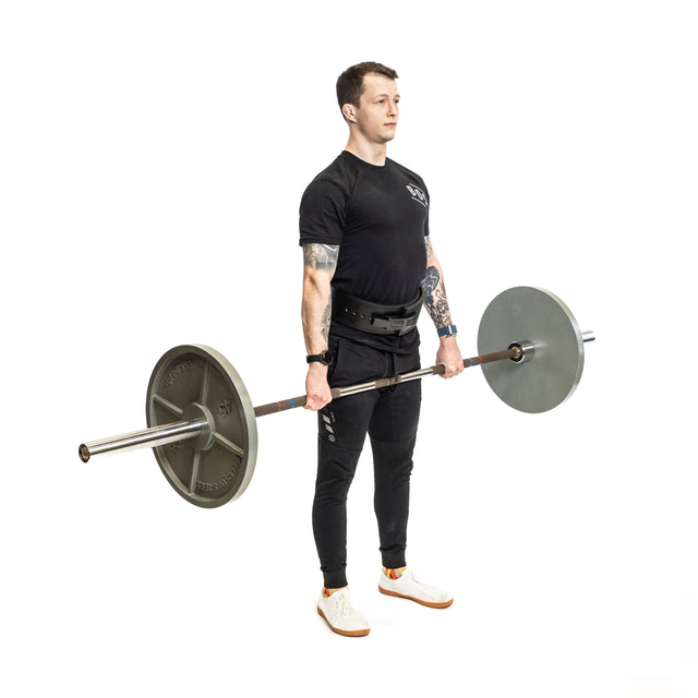 A man in black athletic wear and a Bells of Steel Lever Belt holds a loaded barbell at thigh height, preparing for or finishing a deadlift against a plain white background.