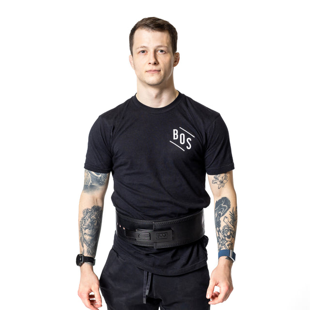 A man with short hair and tattoos on both arms stands facing forward, wearing a black "BOS" t-shirt, a Bells of Steel Lever Belt, and smartwatches. He has a neutral expression against a white background.