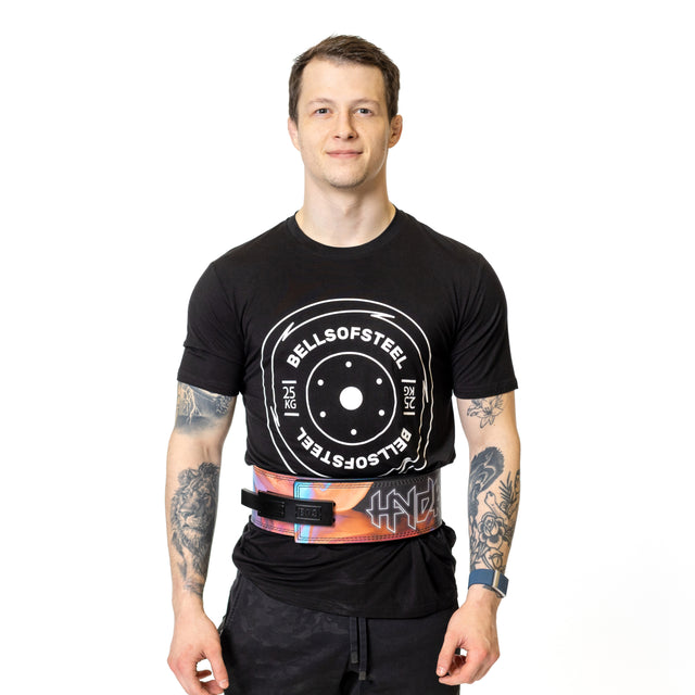 A man with short hair, visible arm tattoos, and black shorts stands facing forward and smiling. He wears a black Bells of Steel t-shirt and the Bells of Steel Lever Belt for powerlifting, posed against a white background.