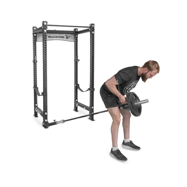 A man in a black shirt and shorts uses the Bells of Steel Landmine Rack Attachment on a power rack to perform a barbell row, seamlessly incorporating rotational core exercises into his strength workout.