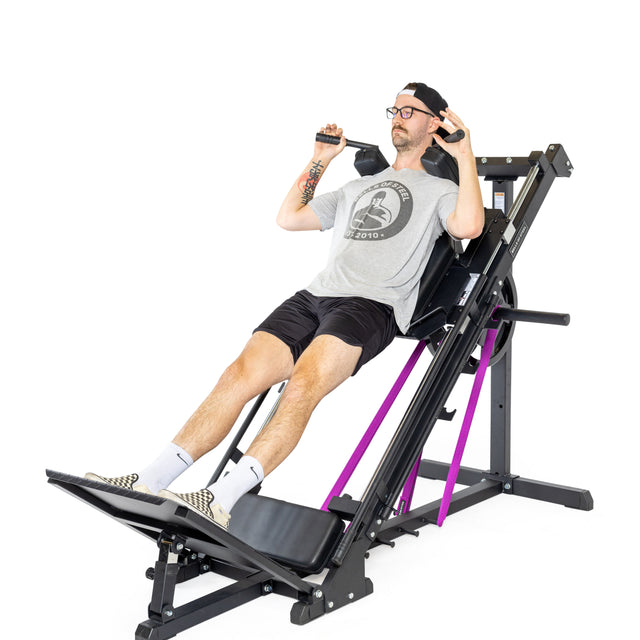 A person in glasses, gray t-shirt, black shorts, and a cap uses the Bells of Steel Leg Press Hack Squat Machine with a black frame and purple accents in their home gym, pushing with their legs while gripping the handles.