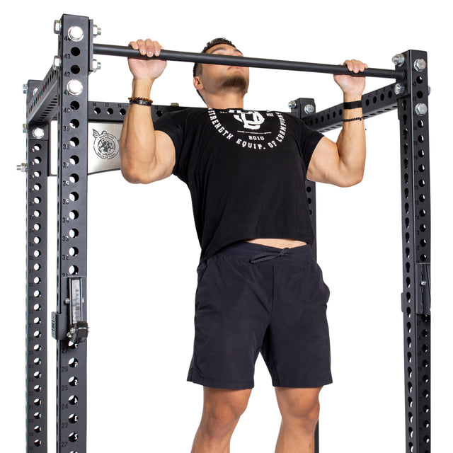 A man in a black t-shirt and shorts does a pull-up on the Bells of Steel Manticore Four Post Power Rack (Prebuilt, 3" x 3", 1" Holes) in his home gym, gripping the bar with both hands and raising his chin above it.