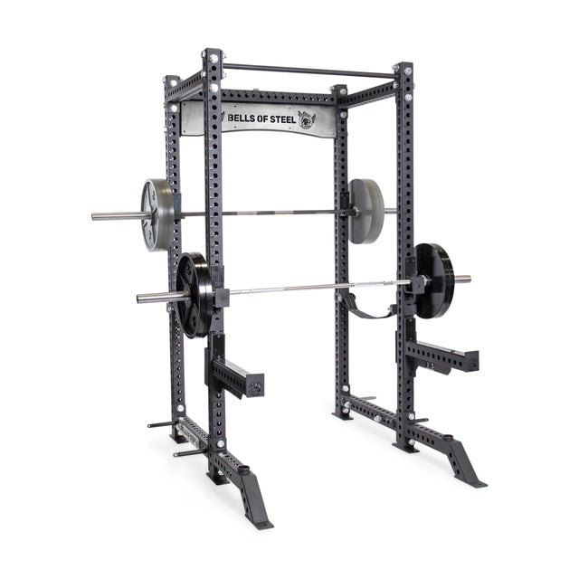 A black Bells of Steel Manticore Four Post Power Rack - Prebuilt (3" x 3", 1" Holes) with safety bars, two loaded barbells, and weight plates on each side stands on a white background, ideal for any home gym.