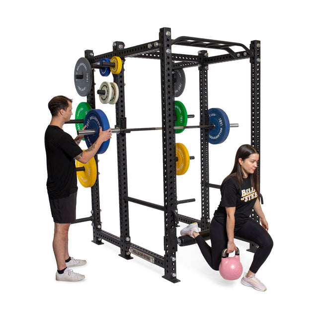 A man adjusts weights on the Bells of Steel Manticore Six Post Power Rack, which features a 1,000-pound capacity and colorful modular components. Nearby, a woman confidently performs a Bulgarian split squat with a pink kettlebell. Both wear sleek athletic clothing.