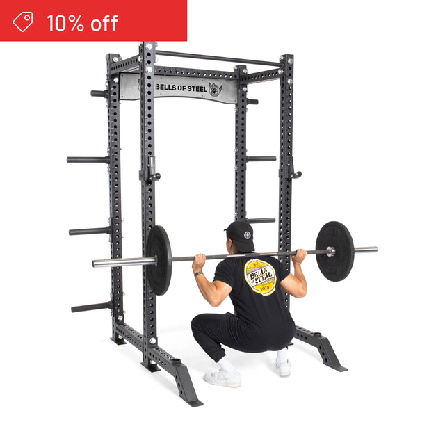 Dressed in black workout gear and a cap, a person does barbell squats inside the Bells of Steel Manticore Collegiate Power Rack Builder (3" x 3", 1" Holes), while a red "10% off" discount banner appears in the top left corner.