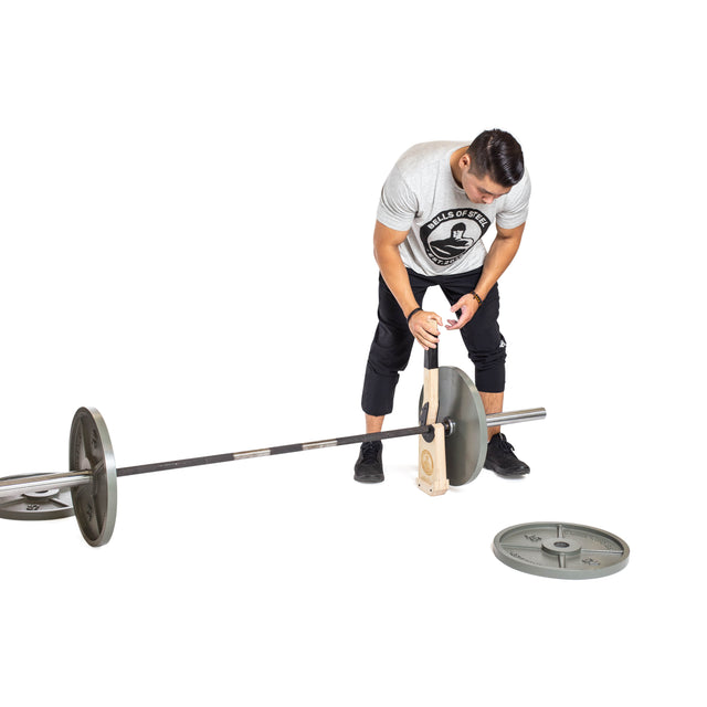 A person in a gray t-shirt and black pants uses the Bells of Steel Meat Hook Deadlift Jack to load a weight plate onto a barbell, with another plate on the floor nearby, all set against a white background.