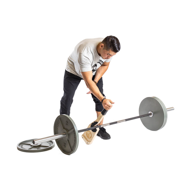 A man in a gray shirt and black pants uses the Bells of Steel Meat Hook Deadlift Jack to lift one end of a loaded barbell for easy plate changes. The scene has a plain white background.