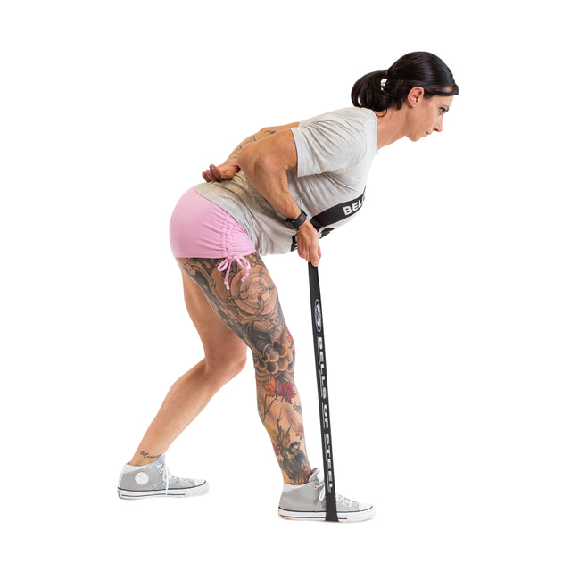 A woman wearing a gray t-shirt and sneakers uses Bells of Steel Mini Bands anchored under her feet for an exercise, displaying her left leg tattoos and highlighting these popular gym accessories.