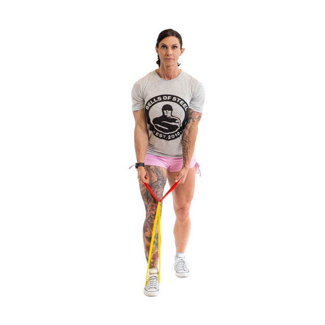 A woman in a gray t-shirt and pink shorts stands on Bells of Steel Mini Bands, holding the handles as she prepares to exercise. She has visible tattoos on her right leg and arm and looks forward against a white background.