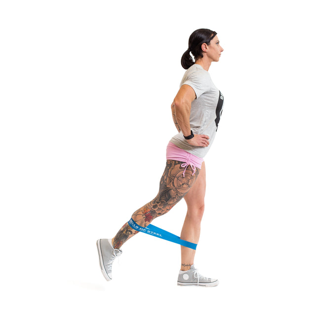 A woman in a gray shirt and pink shorts performs a leg extension, standing on one leg while extending the other back, using Bells of Steel Mini Bands around her ankles.