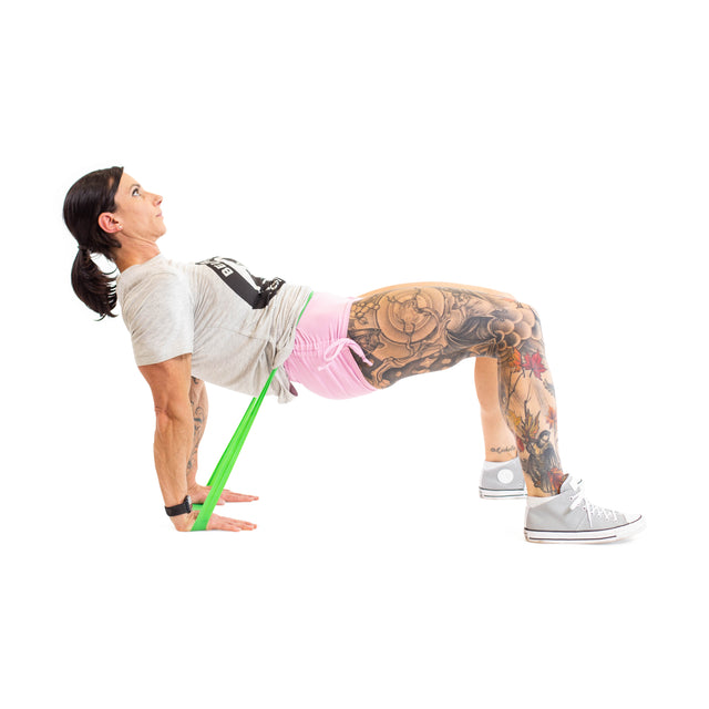 A woman performs a hip thrust using Bells of Steel Mini Bands to increase intensity, supporting herself with her hands and feet on the ground, hips raised, against a white background. Tattoos are visible on her legs.