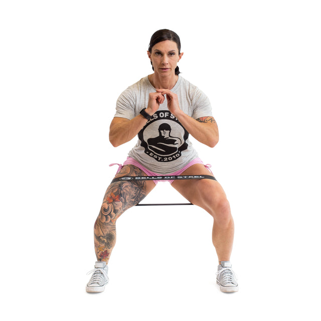 A woman in a gray T-shirt and shorts performs a squat on a white background, using Bells of Steel Mini Bands as resistance around her thighs, with her hands clasped in front of her chest.