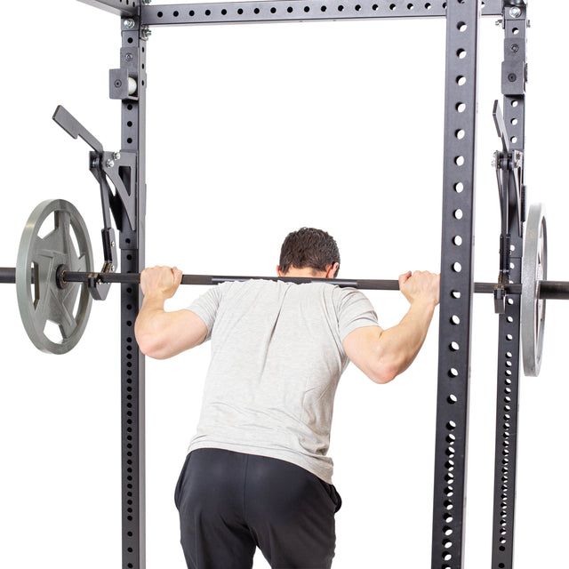 A person in a gray shirt and black pants is squatting inside a squat rack equipped with the Bells of Steel Monolift Rack Attachment, holding a weighted barbell on their shoulders. The scene has a white background.